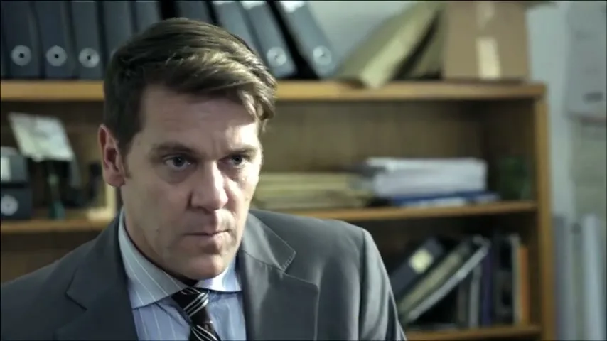 A man in his 30s, wearing a suit and tie, looks up from his desk with a serious expression in an office.