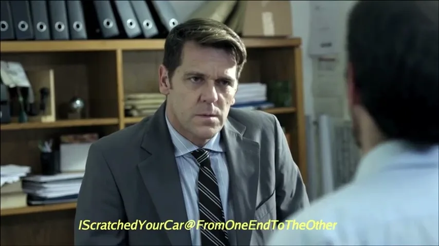 A man in a suit and tie sits at a desk, looking intently at another man whose back is to the camera, in an office.
