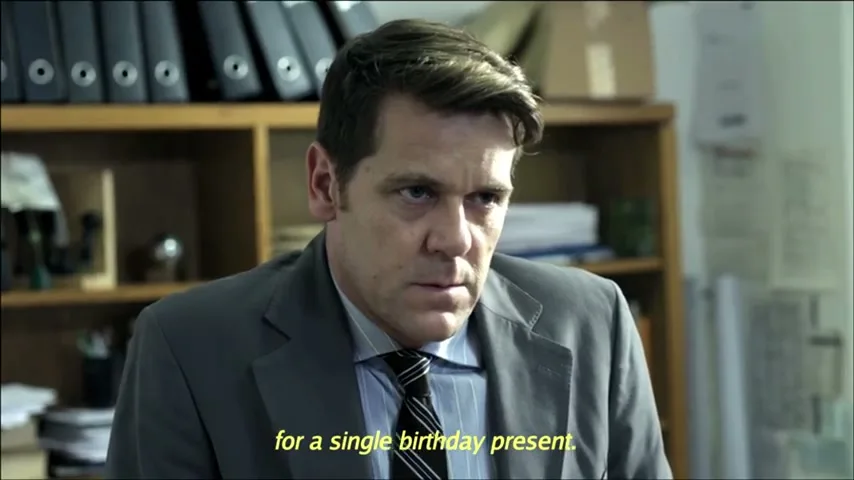 A man in his 40s, wearing a suit and tie, sits at a desk in an office, looking intently at someone off-camera.