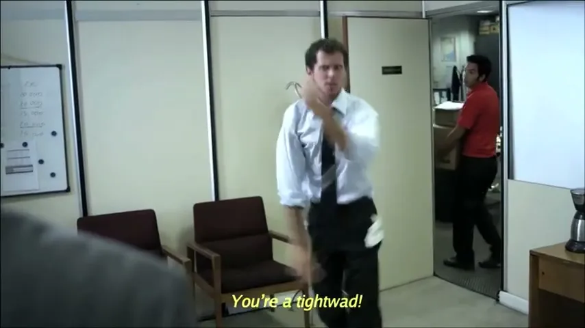 A man in a white shirt and tie dances wildly in an office, celebrating the departure of a colleague, while another man carries boxes in the background.