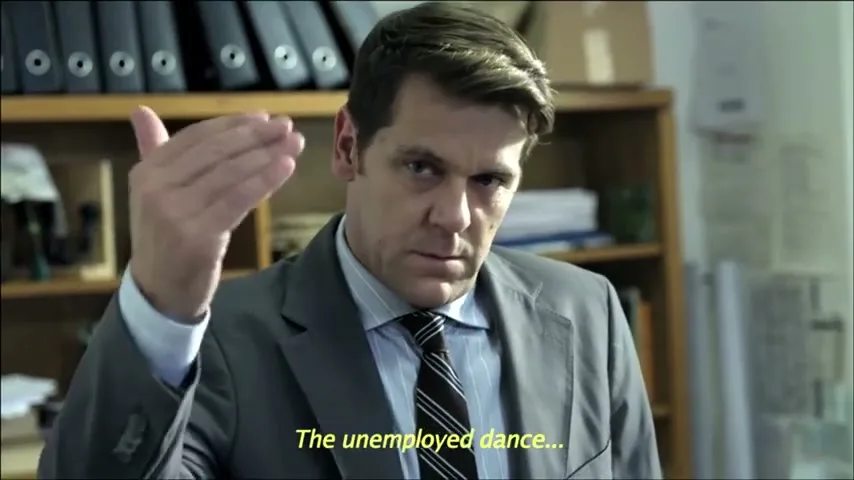 A man in a suit and tie sits at a desk in an office, looking at the camera and making a gesture with his hand.