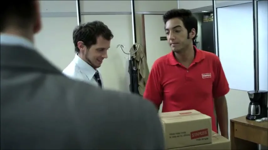 A man in a red shirt delivers Staples boxes to an office worker, while another man in a white shirt and tie looks on, in an office setting.