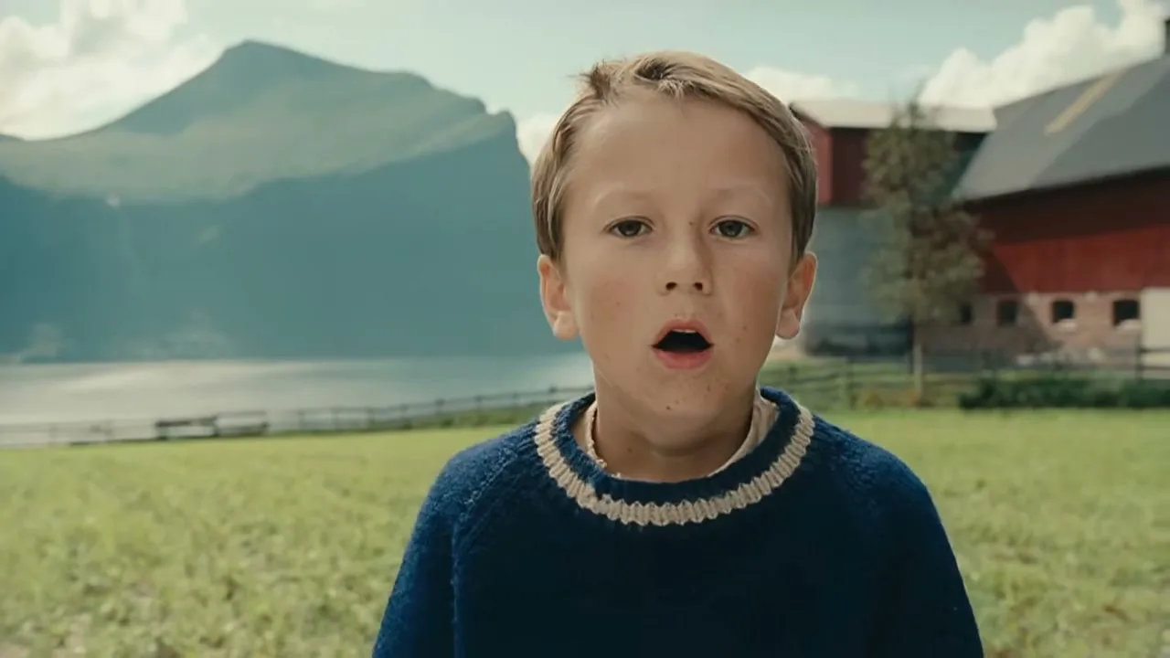 A young boy with light brown hair and a blue sweater looks at the camera with his mouth slightly open in a rural, mountainous landscape.