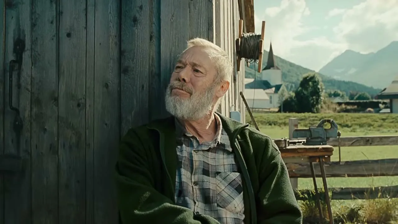 A bearded older man in a green jacket and plaid shirt leans against a wooden barn, looking up thoughtfully on a sunny day in a rural setting.