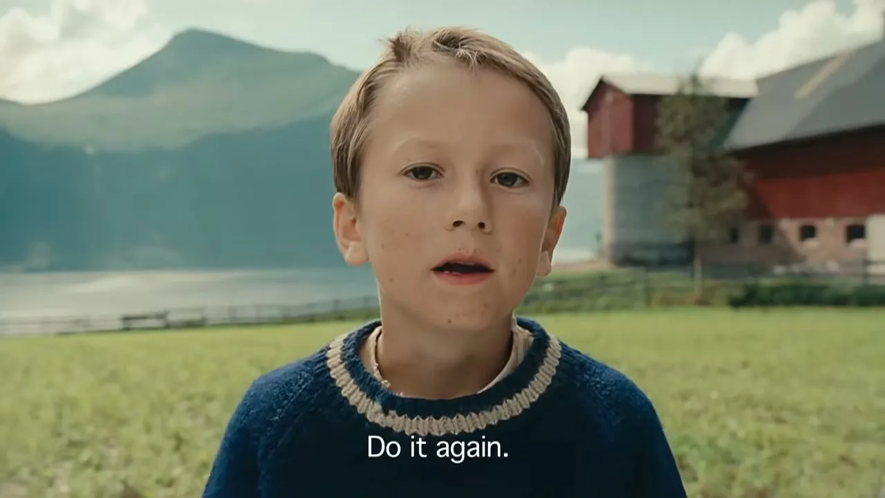 A young boy with blonde hair and a blue sweater looks at the camera, asking for a magic trick to be repeated in a rural, mountainous setting.