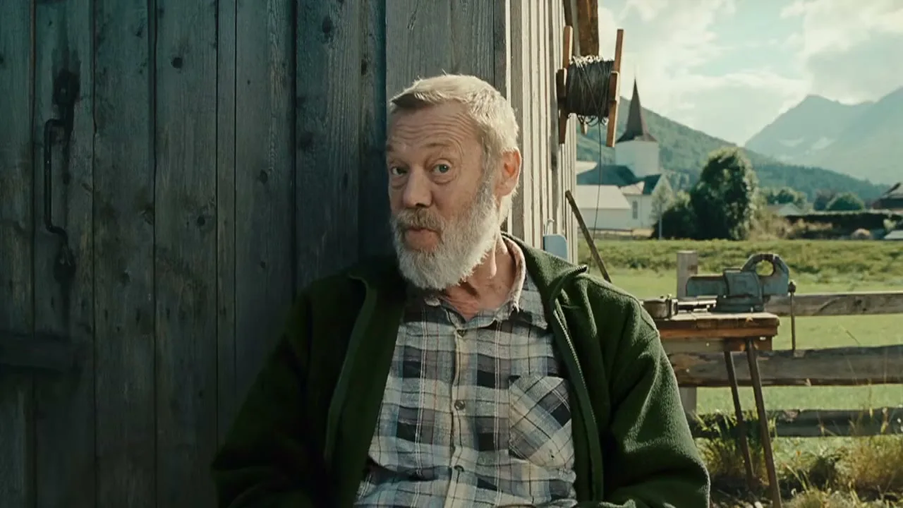 An elderly man with a white beard drinks from a mug, then looks at the camera and smiles, sitting outside a wooden building in a rural setting.