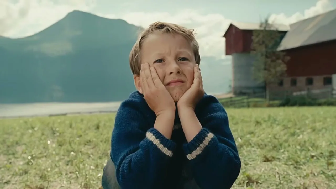 A young boy with blonde hair and a blue sweater sits in a grassy field, looking up with a pleading expression, with mountains and a barn in the background.