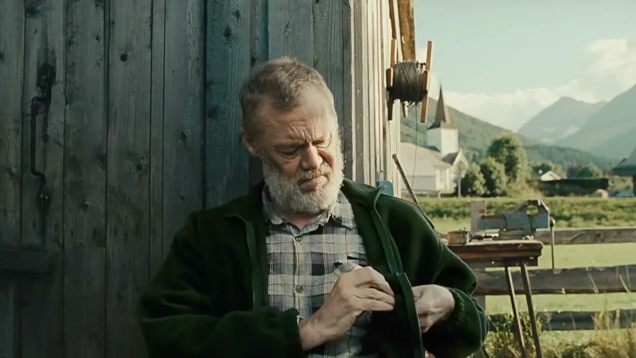 An old man with a white beard performs a magic trick with a spoon, then puts it in his pocket, outside a wooden shed in a rural valley.
