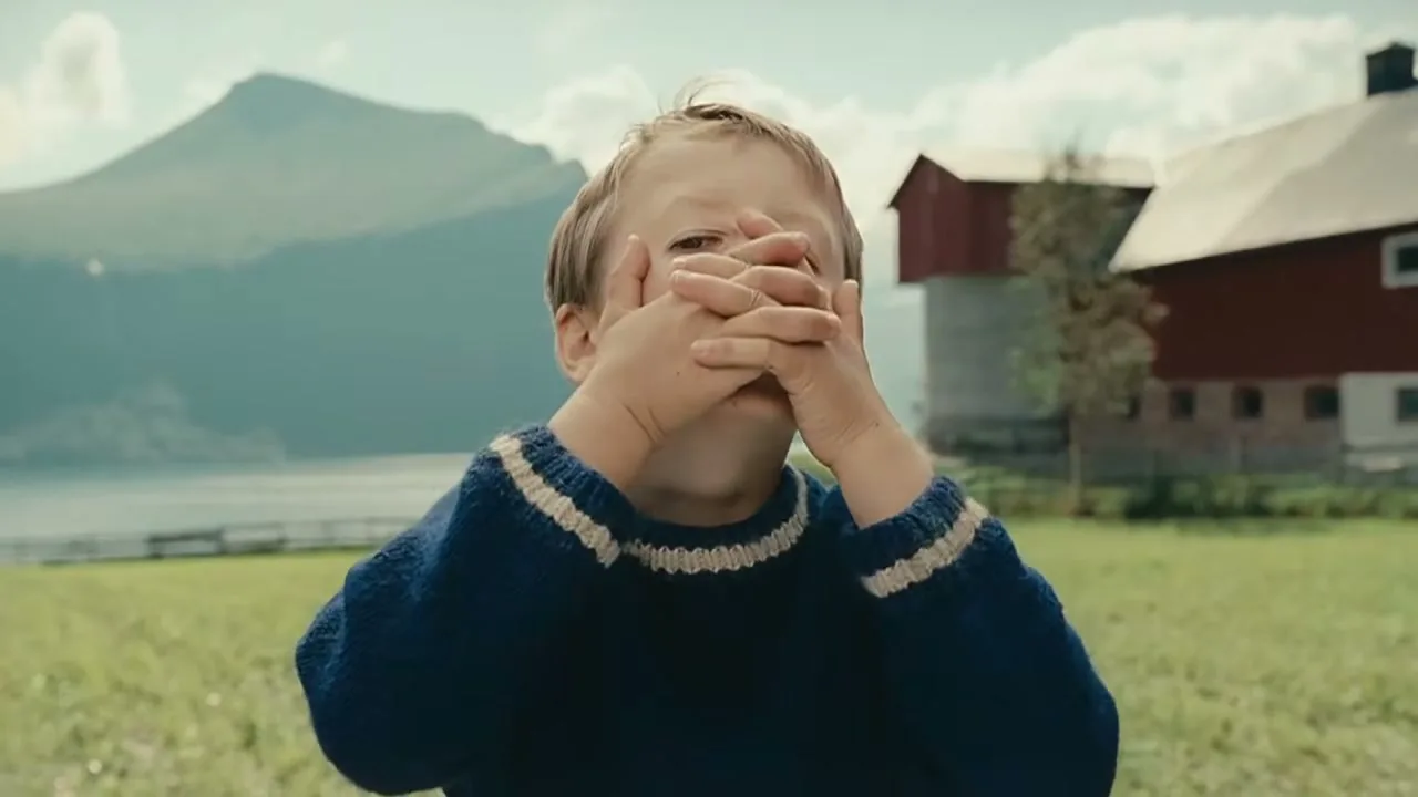 A young boy in a blue sweater covers his eyes, then opens them, looking directly at the camera in a scenic outdoor setting with mountains and a lake.