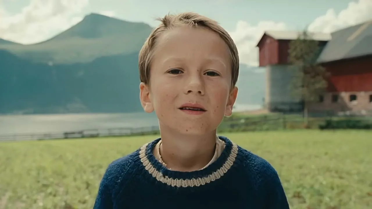 A young boy with blonde hair and a blue sweater looks directly at the camera in a green field with mountains and a red barn in the background.