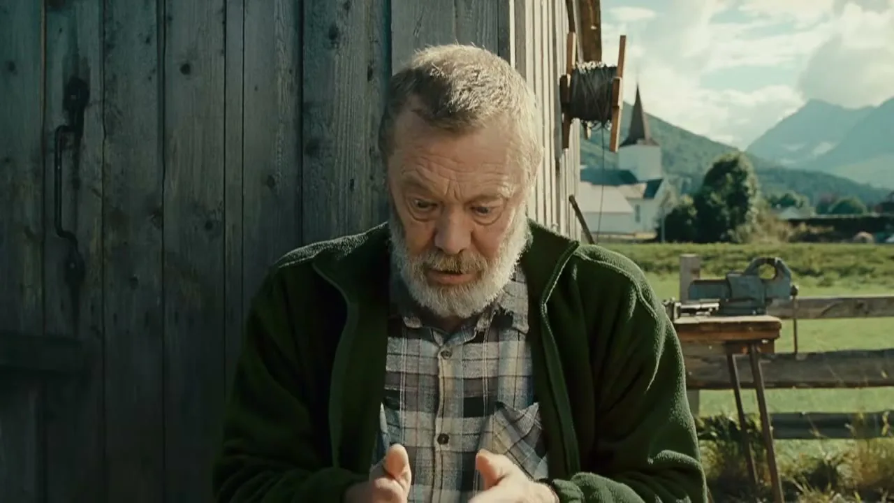An older man with a white beard and green jacket looks surprised and claps his hands together in front of a wooden barn with mountains in the background.