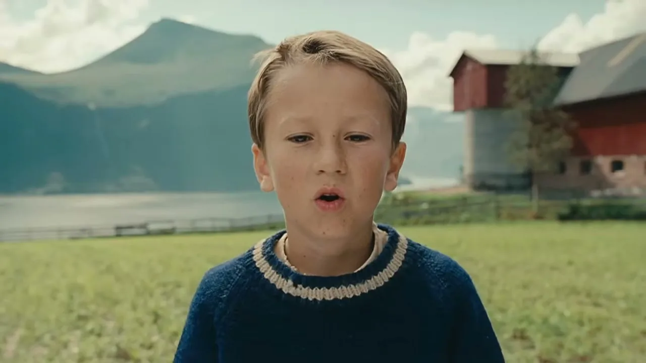 A young boy with blonde hair and a blue sweater stands in a green field, looking directly at the camera with a slightly open mouth, with mountains and a red barn in the background.