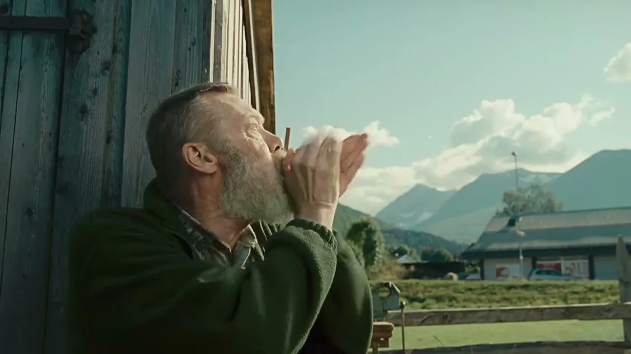 An older man with a white beard blows into his hands, then looks up at a plane flying over a mountainous landscape, and smiles.