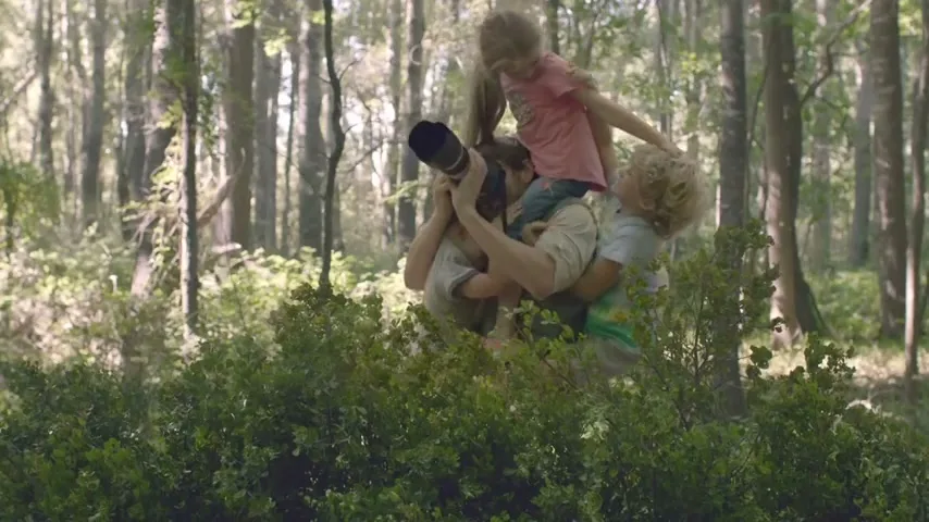 A father with a camera on his head carries two young children on his shoulders and back, peeking over green bushes in a forest.