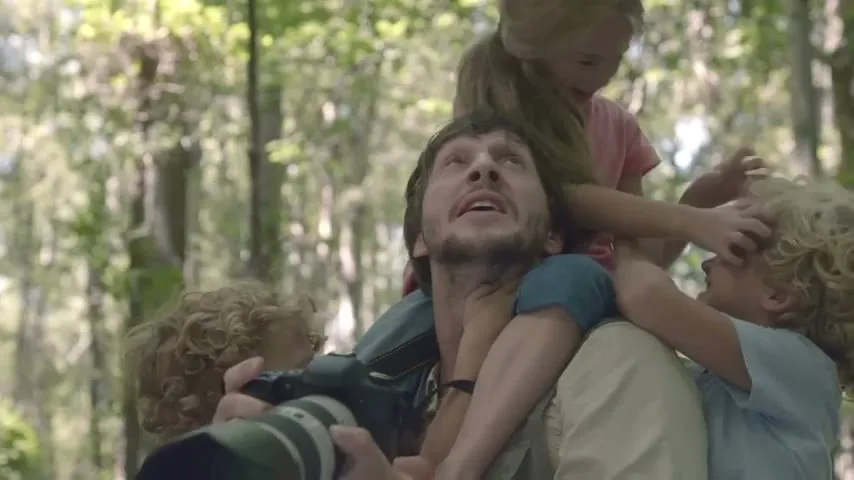 A father with a camera looks up, while two young children climb on his shoulders and head in a sun-dappled forest.