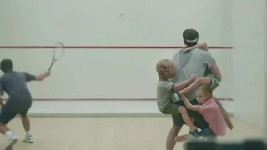 A man plays squash while another man is playfully burdened by three children clinging to him in a squash court.