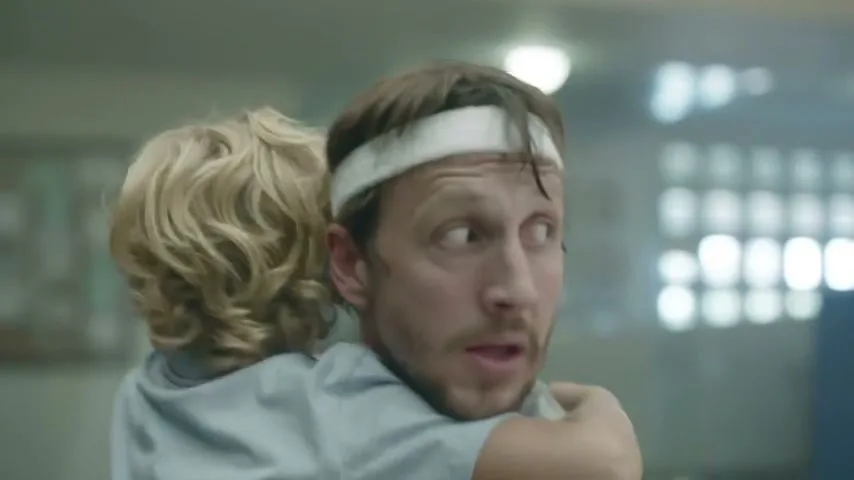A man with a white headband holds a young child in a light blue shirt, looking around nervously in an interior setting.