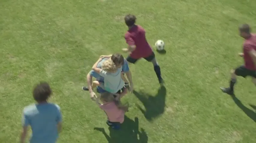 A father is embraced by his three children on a sunny green soccer field, while other players run around them.