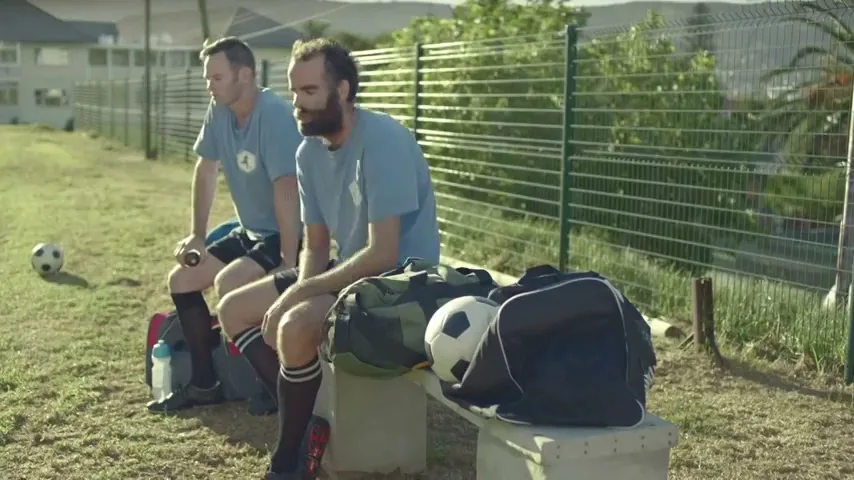 Two adult men in light blue soccer jerseys and black shorts sit on a bench next to soccer bags and a ball on a grassy field during the day.