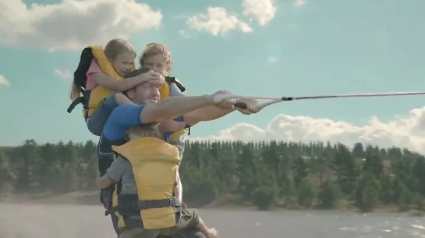 A father water skis while carrying two young children on his back and shoulders, all wearing yellow life vests, on a lake under a blue sky.