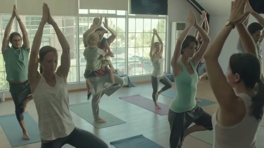 A diverse group of adults and one child practice yoga in a bright studio with large windows overlooking a parking lot.