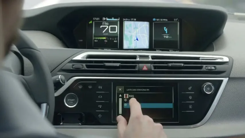 A person's hand interacts with the touchscreen display of a car's dashboard, showing navigation and radio controls while driving.