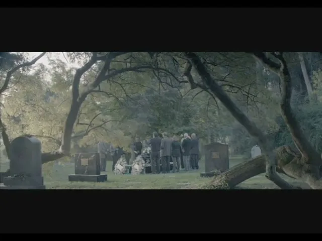 A group of people dressed in dark clothing gather around a casket at a graveside funeral in a cemetery, surrounded by trees.