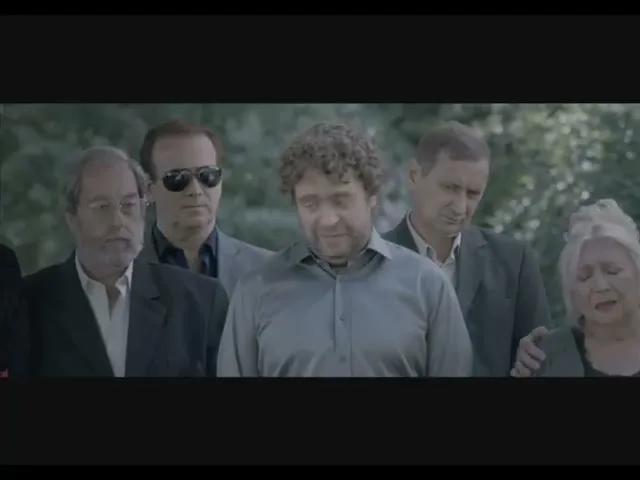A group of mourners, including a man with curly hair, stand solemnly at an outdoor funeral, looking downcast and grieving.