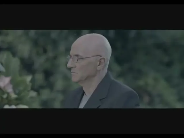 A bald, older man in glasses and a dark suit stands in profile, looking off-screen with a neutral expression, outdoors amidst blurred greenery.