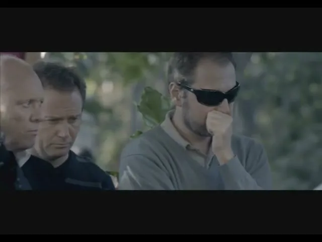 A man in sunglasses covers his mouth, looking distraught, surrounded by other men at an outdoor gathering.