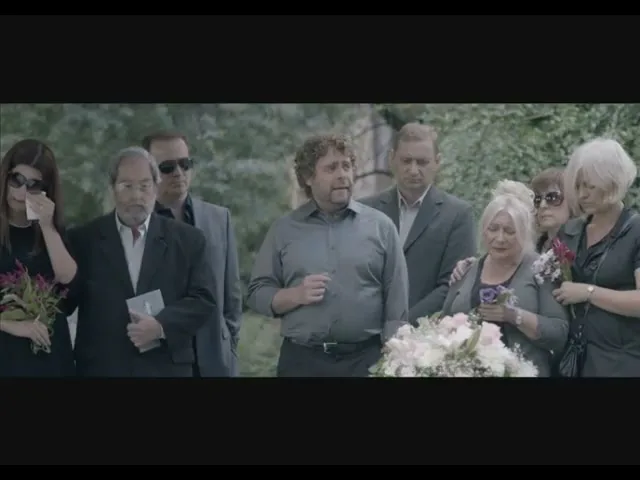 A group of people, dressed in dark clothing, stand around a flower-covered casket at an outdoor funeral, with one man speaking animatedly.