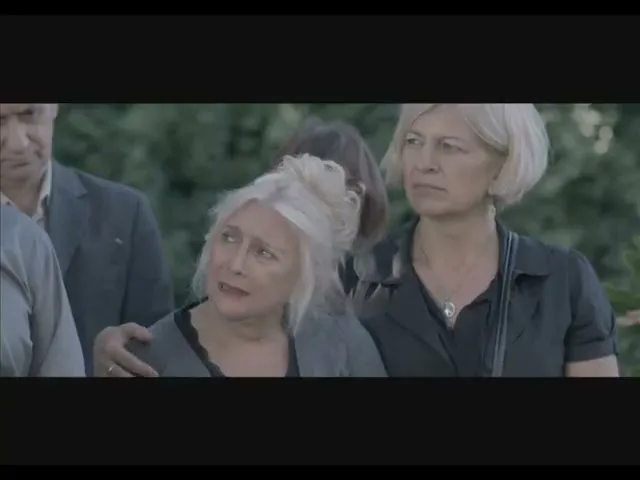 A group of mourners, including an older woman with blonde hair, stands somberly at an outdoor funeral, with a man's hand on her shoulder.