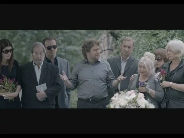 A man with curly hair gestures animatedly while speaking to a group of mourners at a funeral, with a casket and flowers in the foreground.