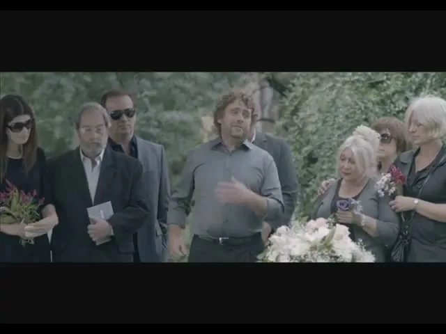 A man with curly hair speaks animatedly at a funeral, surrounded by grieving attendees in an outdoor cemetery setting.