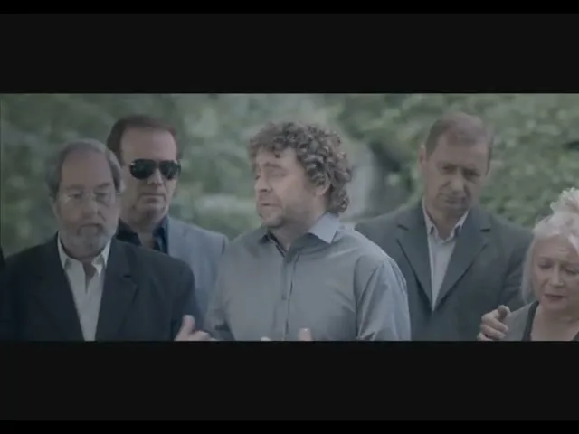 A man with curly hair in a grey shirt speaks animatedly, gesturing with his hands, surrounded by four other people in a somber outdoor setting.