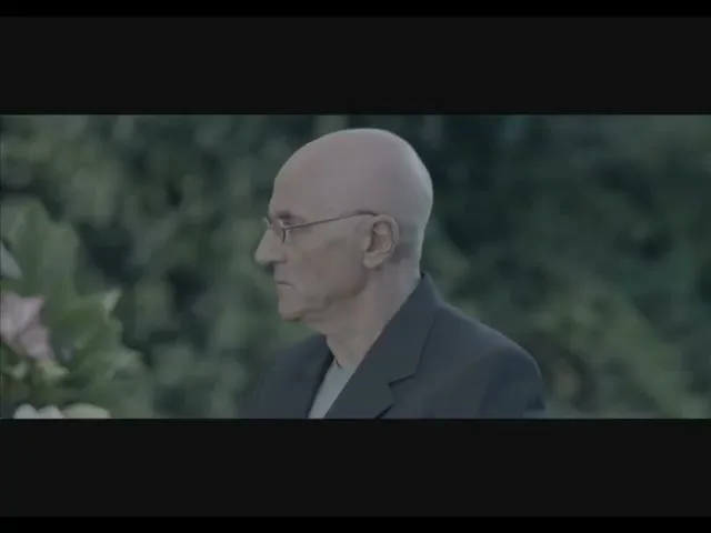 A bald, older man with glasses and a dark suit stands outdoors, looking off-camera with a neutral expression.