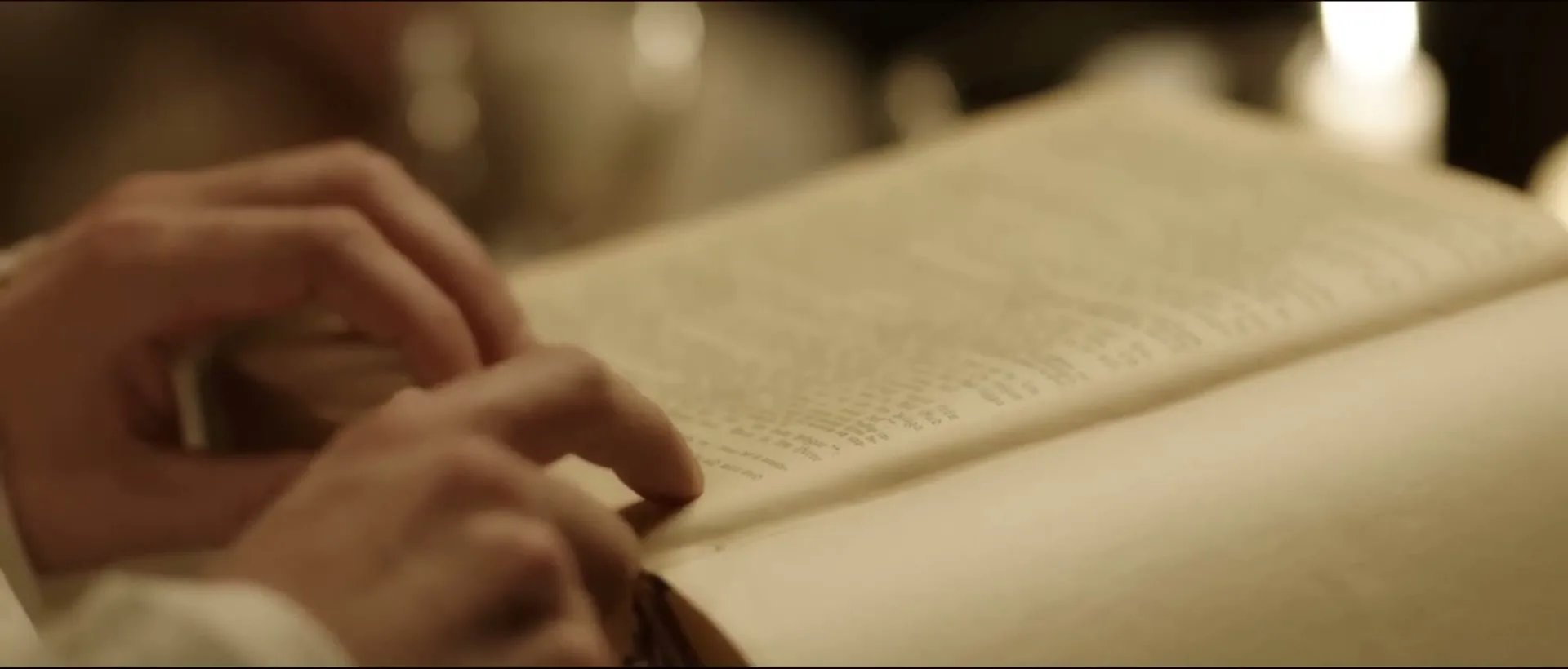 A person's hands are seen turning the pages of an old, thick book, possibly a bible, in a dimly lit room.