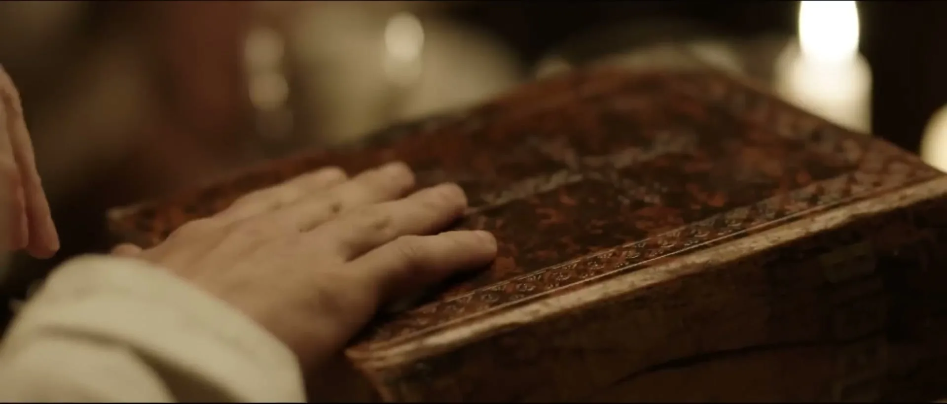 A person's hand in a white sleeve closes an old, ornate book on a dark table, with a candle in the background.