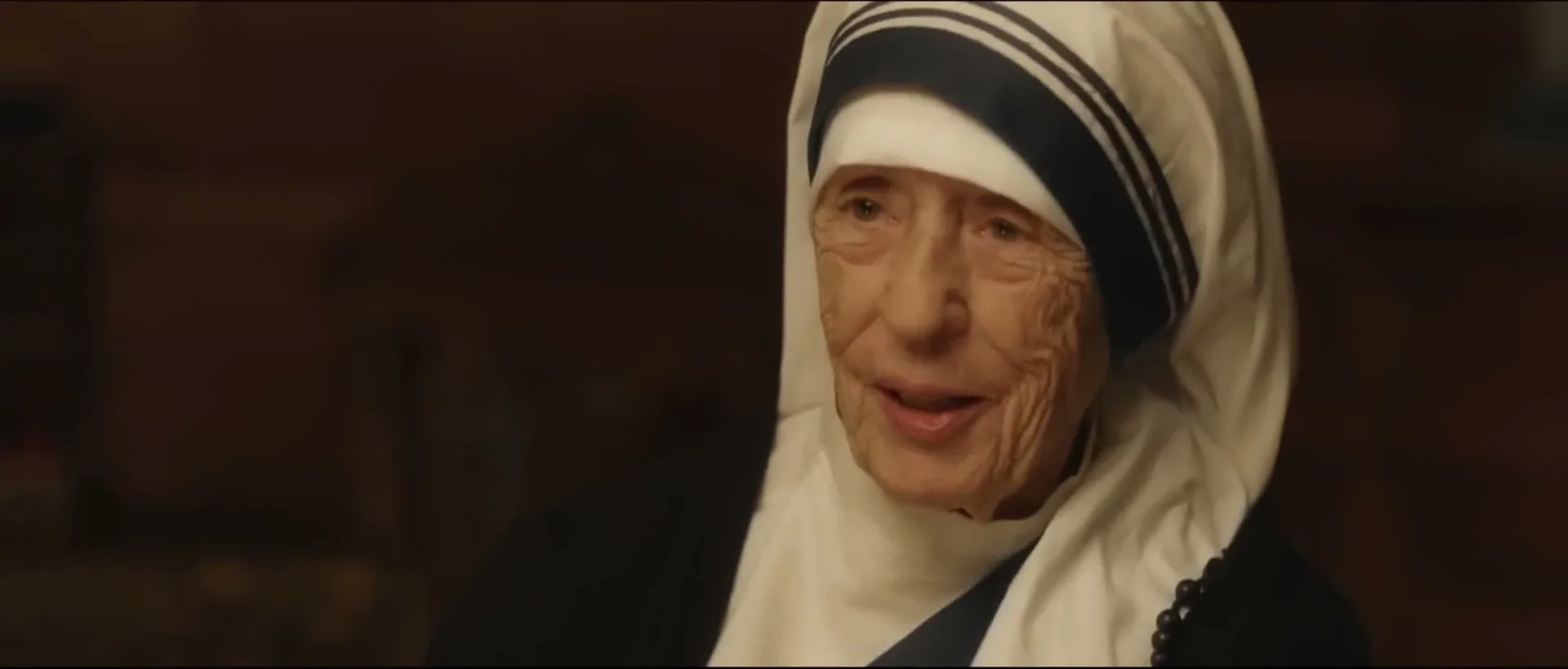 Mother Teresa, an elderly woman in a white and blue habit, smiles gently while speaking in a dimly lit interior.