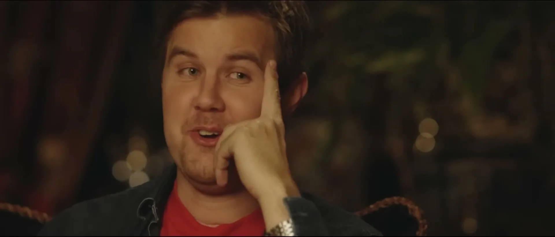 A man in his 30s, wearing a red t-shirt and blue jacket, sits with his hand on his face, talking and smiling in a dimly lit room.