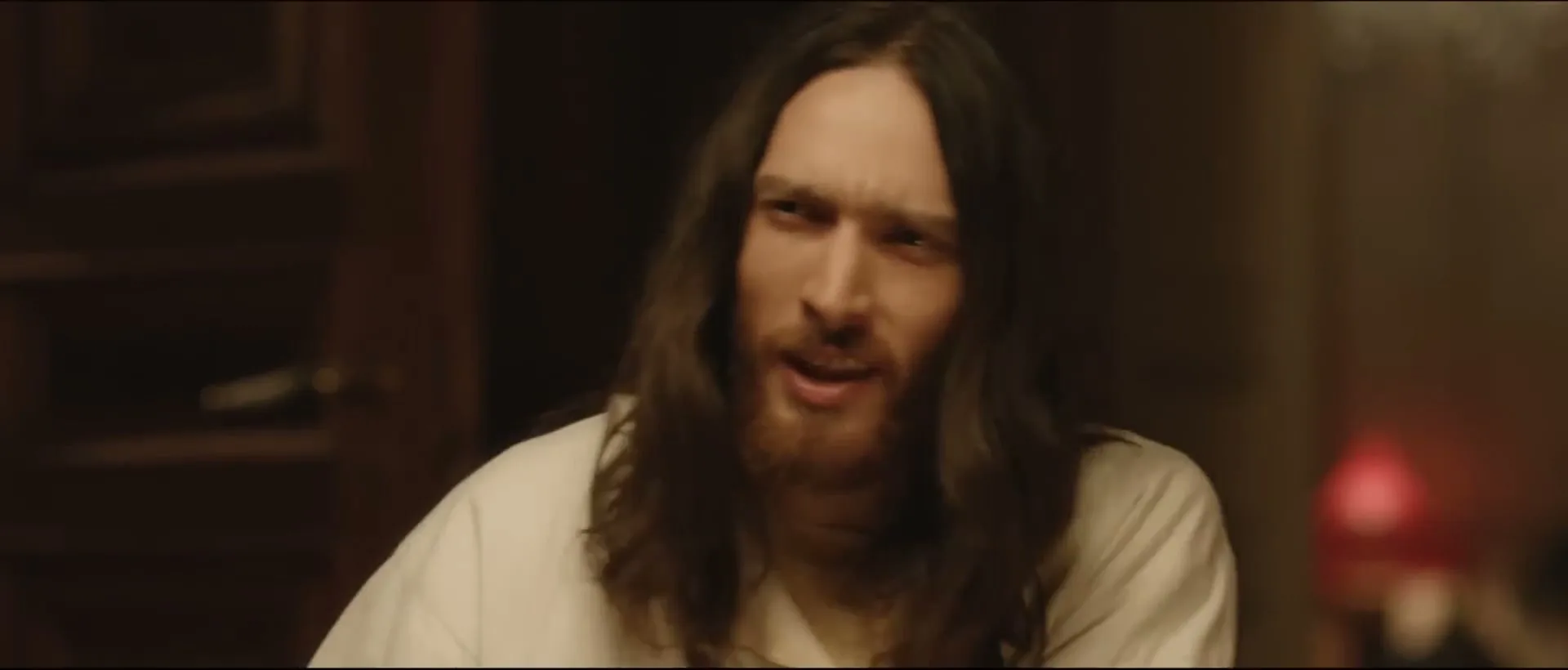 A young man with long brown hair and a beard, wearing a white shirt, smiles and talks in a dimly lit room.