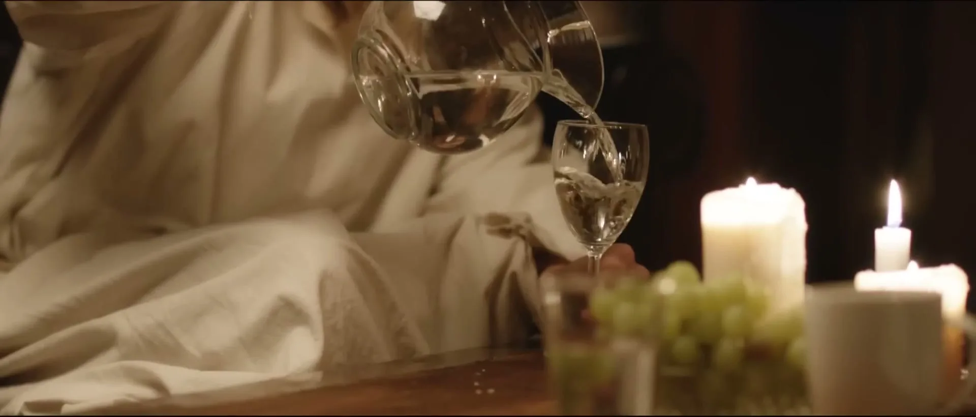 A person in a white robe pours water from a glass pitcher into a wine glass on a candlelit table with grapes.