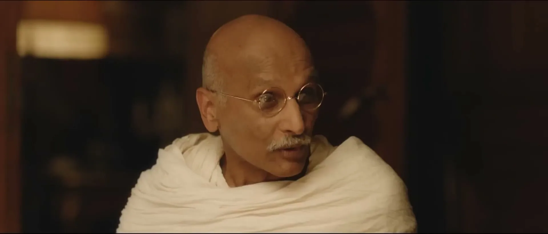 An older man with a mustache and glasses, dressed in a white shawl, speaks in a dimly lit room.