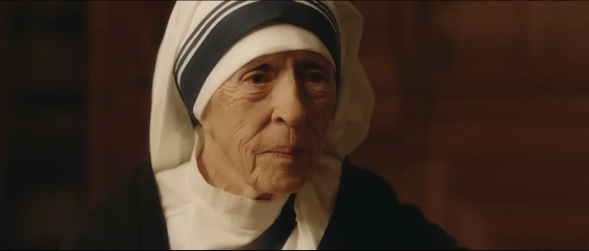 An elderly woman, resembling Mother Teresa, with a white and blue habit, looks forward with a gentle expression in a dimly lit interior.