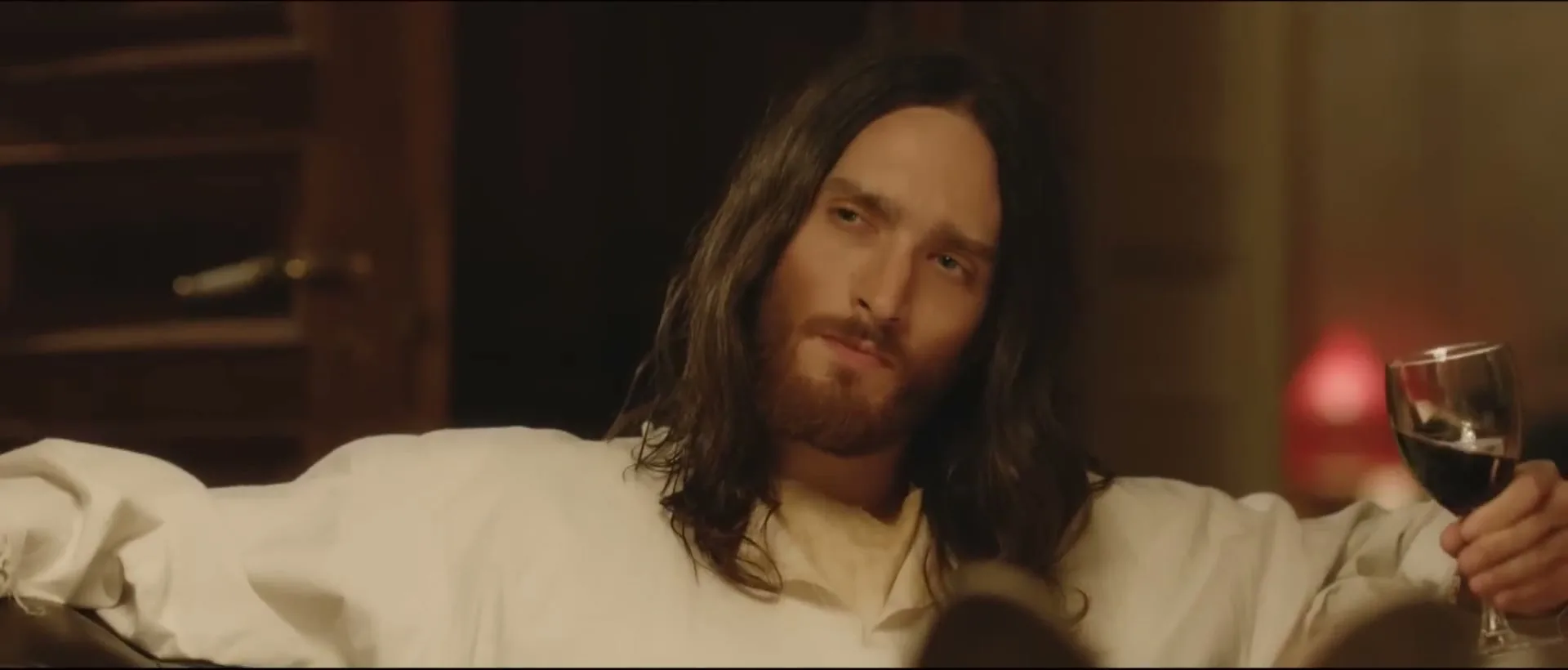 A man with long dark hair and a beard, dressed in a white robe, sips red wine and looks thoughtfully at the camera in a dimly lit room.