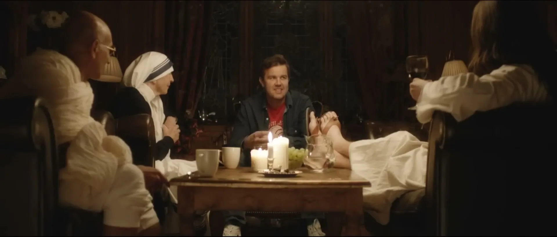 A man with a beard sits at a candlelit table with three other people, one of whom has their feet on the table, in a dimly lit room.