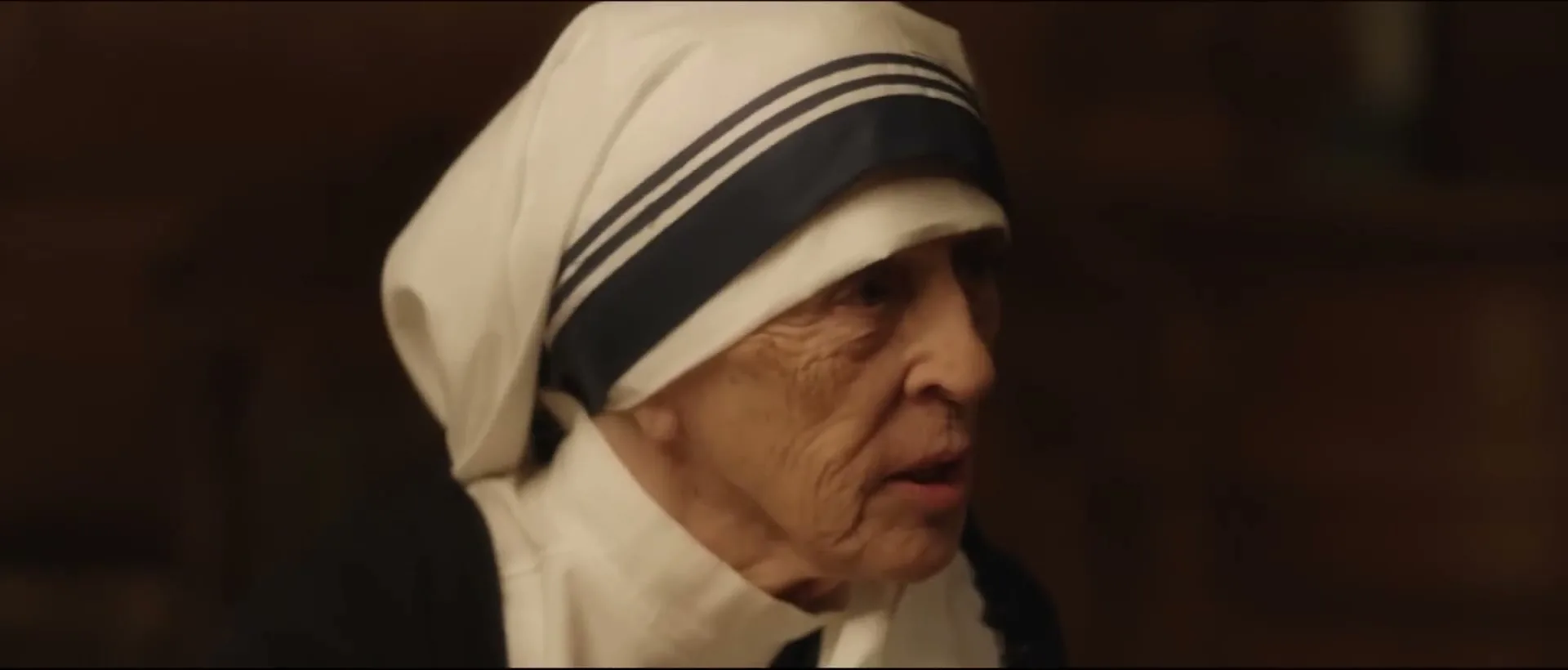 An elderly woman, resembling Mother Teresa, wearing a white habit with blue stripes, speaks with a serious expression in a dimly lit interior.