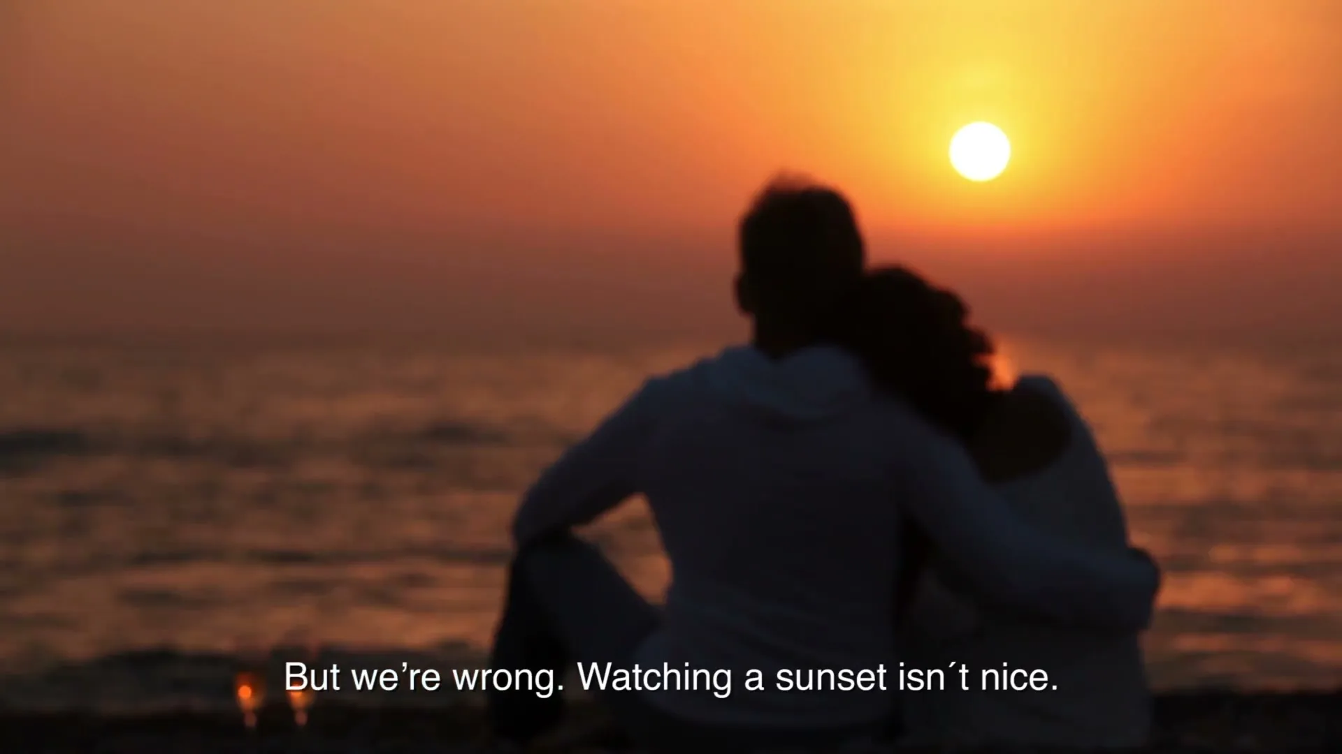 A couple sits on a beach, silhouetted against a vibrant sunset over the ocean, with two champagne glasses in the foreground.