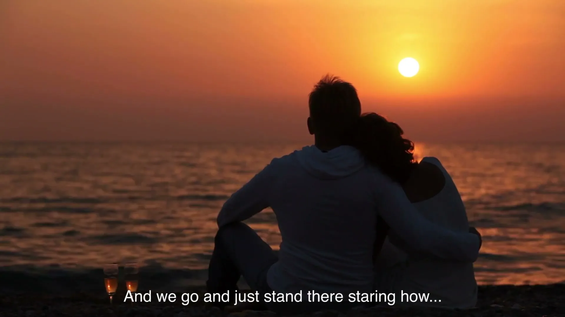 A couple sits on a beach, silhouetted against a vibrant orange sunset over the ocean, with two champagne glasses beside them.