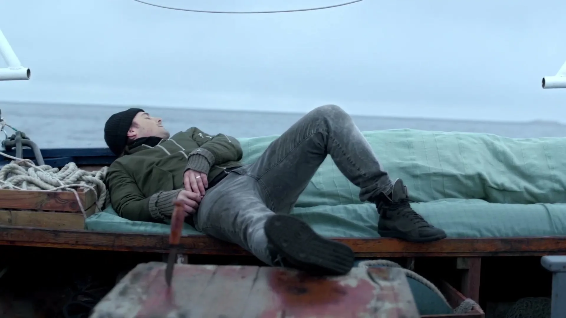A man in a black beanie and green jacket lies on a boat deck, looking up at the sky, with the ocean in the background.